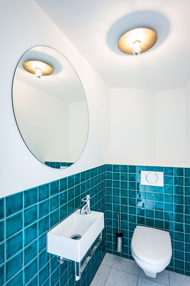 wc-mit-kleinem-waschbecken-catalano-und-rundem-spiegel-sowie-lehni-aluminium-leuchte-und-blaugruenen-handgefertigten-kacheln