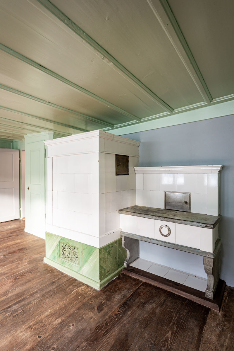 restaurierter-kachelofen-mit-sitzbank-mit-weissen-kacheln-und-gruen-marmoriertem-sockel-aus-der-zeit-um-1900-im-gruenen-zimmer